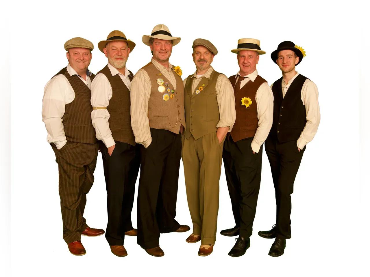 Sechs Männer von der Band Sunshine Brass mit beigen Hüten, cremfarbenen Hemden, Weste und Anzugshose, die eine Sonnenblume angesteckt haben.