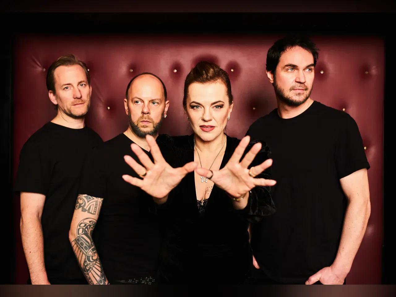 Die Band von DIE HAPPY mit drei Männern und einer Frau, die die Hände nach vorne streckt, alle in schwarzen T-Shirts vor einem roten Polster.