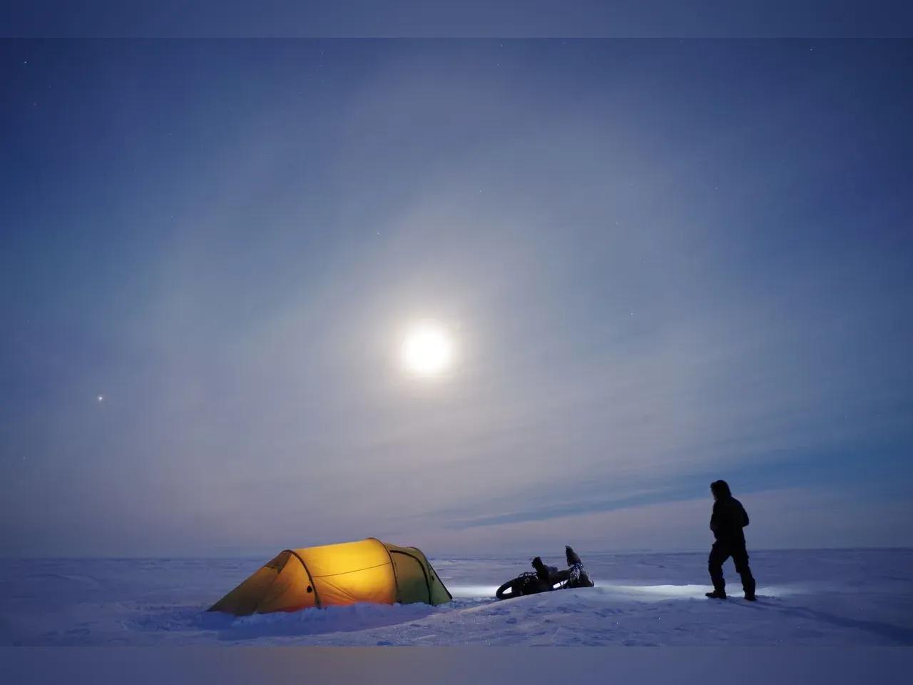 Schneedecke unter blauem Himmel, darauf ein erleuchtetes Zelt, ein Schneemobil und Richard Löwenherz im Schneeanzug