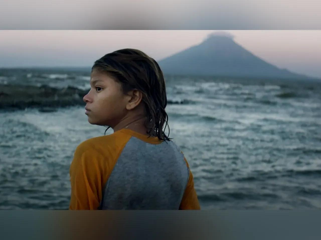 Filmszene aus "La hija de todas las rabias": Mädchen steht mit nassen Haaren vor dem Meer, ein Vulkan im Hintergrund